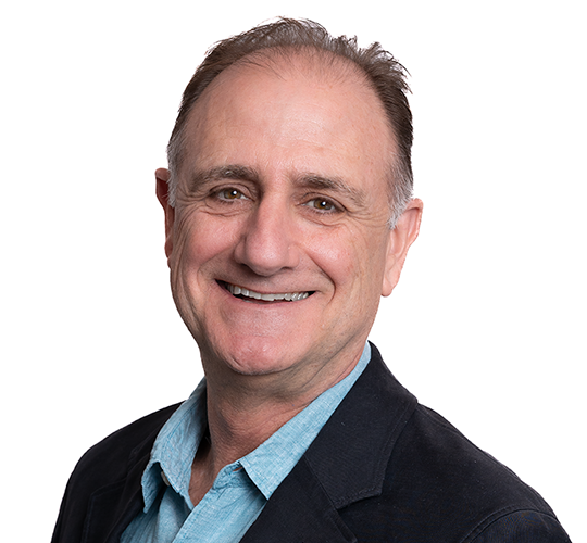 A middle-aged man smiling, wearing a blue collared shirt and a dark blazer, against a white background.
